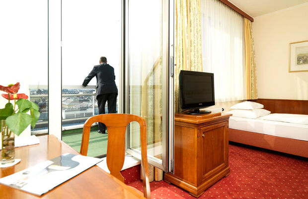 Ein Einzelzimmer mit einem Bett, einen Schreibtisch und einem Fernseher. Auf dem Balkon steht ein Mann.