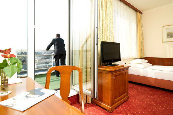 Ein Einzelzimmer mit einem Bett, einen Schreibtisch und einem Fernseher. Auf dem Balkon steht ein Mann.